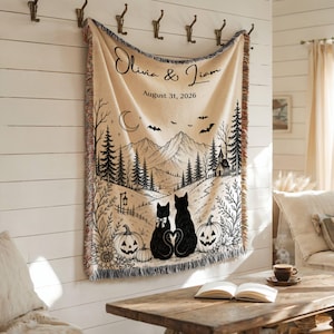May include: A cream-colored woven throw blanket featuring a black ink-style illustration of two cats with tails forming a heart, pumpkins, a mountain landscape, pine trees, bats, and a crescent moon. The text reads Olivia & Liam August 31, 2026.