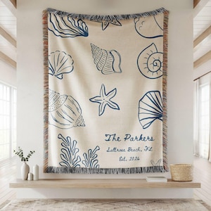 May include: A cream-colored woven throw blanket featuring a dark blue line-art pattern of various seashells, starfish, and coral. The text reads The Parkers, Saltrose Beach, FL, Est. 2026. The blanket has a multi-colored fringed edge on all sides.