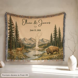 May include: A woven throw blanket featuring a landscape with a river, mountains, pine trees, a deer, and a bison. The text reads Olivia & James and June 15, 2026, with a heart icon. The blanket has a fringed edge and a beige background color.