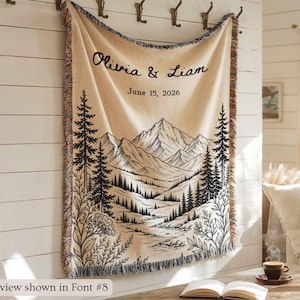 Personalized mountain woven blanket with custom couple names and date, featuring forest trees and rustic mountain landscape art, styled as cozy cabin wall decor for wedding, engagement, or anniversary gift.