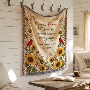 May include: A cream-colored woven throw blanket featuring a sunflower and red cardinal bird border. The text reads: Those we love don't go away. They walk beside us every day. LAUREN GARCIA PARKER 1964-2023. The blanket has a fringed edge.