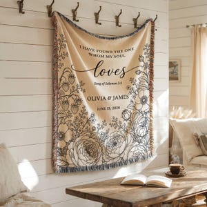 May include: A cream-colored woven throw blanket featuring black line art of flowers and strawberries. The text reads: I HAVE FOUND THE ONE WHOM MY SOUL loves, Song of Solomon 3:4, OLIVIA & JAMES, JUNE 15, 2026. The blanket has fringed edges.