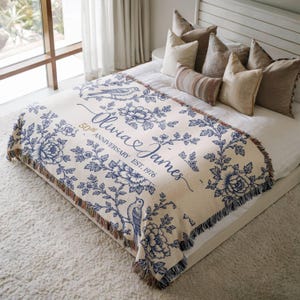 May include: A cream-colored woven throw blanket featuring a blue floral and bird pattern. The text reads Olivia & James, 50th Anniversary Est. 1976. The blanket has a fringed edge and is draped over a bed with several neutral-toned decorative pillows.