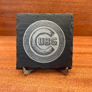 Op de afbeelding: Een vierkante zwarte leistenen onderzetter met een in het midden lasergegraveerd Chicago Cubs logo. Het logo bestaat uit een grote letter C met het woord CUBS horizontaal in het midden geschreven. De onderzetter heeft een ruwe natuursteenstructuur.