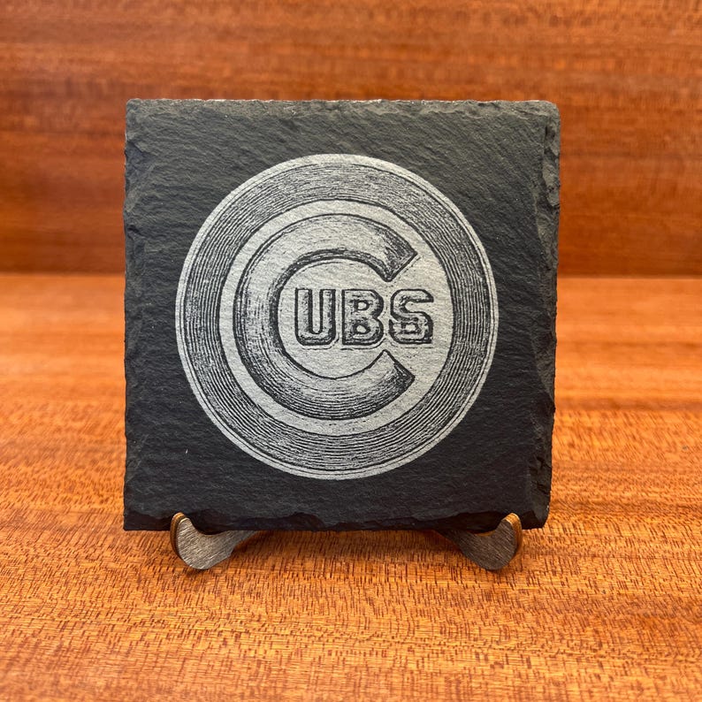 Op de afbeelding: Een vierkante zwarte leistenen onderzetter met een in het midden lasergegraveerd Chicago Cubs logo. Het logo bestaat uit een grote letter C met het woord CUBS horizontaal in het midden geschreven. De onderzetter heeft een ruwe natuursteenstructuur.