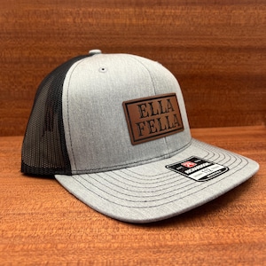 May include: A gray and black trucker hat with a curved brim. The hat features a brown leather patch with the text "ELLA FELLA". The hat has a mesh back and a snap closure.