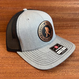 Könnte beinhalten: Eine graumelierte Trucker-Cap mit schwarzem Netzrücken und flachem Schirm. Auf der Vorderseite befindet sich ein runder Aufnäher mit einer kupferfarbenen Bigfoot-Silhouette und dem Text OFFICIAL BIGFOOT SEARCH TEAM. Ein schwarzer Aufkleber zeigt RICHARDSON 112.