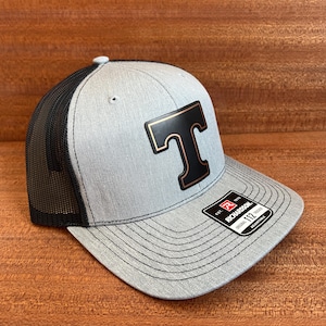 Può includere: Cappellino da camionista grigio e nero con logo "T" nero bordato d'oro. Il cappellino ha un retro in rete e una visiera curva. L'etichetta del marchio Richardson è visibile.