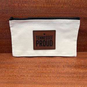 Peut inclure: Une pochette zippée rectangulaire en toile blanc cassé avec un écusson central en similicuir marron. L'écusson présente une petite étoile au-dessus du texte noir en gras "TENNESSEE PROUD" encadré par une bordure rectangulaire sur le tissu.