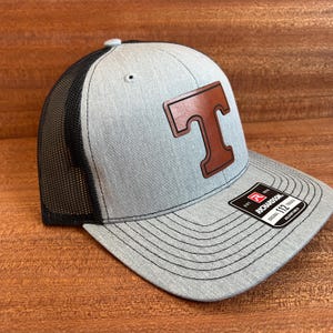 May include: A grey and black trucker cap with a brown leather patch displaying the letter "T". The cap has a curved peak and a black mesh back. The Richardson 112 cap is adjustable.