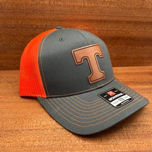 May include: A grey and orange trucker cap with a brown leather "T" patch. The cap has a curved peak and orange mesh back panels. It is adjustable and features a Richardson 112 label. The hat is a popular style.
