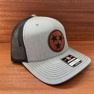 May include: A grey trucker hat with a black mesh back and a brown circular patch on the front featuring three black stars. A black sticker on the brim displays the text Richardson Original 112 Trucker. The hat has a structured crown and a flat brim.
