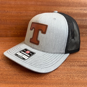 Può includere: Cappellino da camionista grigio e nero con toppa in pelle marrone con una "T". Il cappello ha una visiera curva e un retro in rete. Il cappellino Richardson 112 è regolabile.