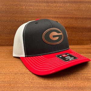 May include: A baseball cap featuring a black front panel with a copper-colored letter G logo, white mesh back panels, and a red brim. A black sticker on the brim displays the text 112 The Original MD-LG with a red R logo and white lettering.