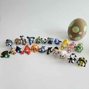 Könnte beinhalten: Eine Sammlung farbenfroher, miniaturisierter Tierfiguren, darunter eine Giraffe, eine Schildkröte und ein Hummer. Ein brauner, eiförmiger Behälter mit grünen Punkten ist ebenfalls vorhanden. Die Figuren sind auf einer weißen Oberfläche angeordnet.
