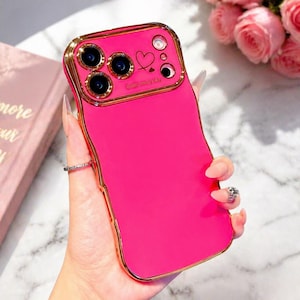 May include: A bright pink phone case with a gold-colored frame. The case has a heart and the words "Smile every day" near the camera lenses. The case is held in a hand with a ring on the finger.
