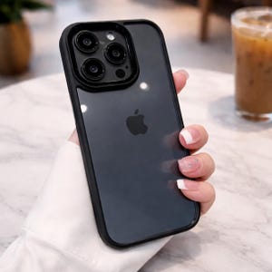 Peut inclure: Une coque de téléphone noire pour iPhone avec un dos transparent et un bord noir. L'étui présente une découpe pour l'appareil photo avec trois lentilles et un flash. Le logo Apple est visible. Le téléphone est tenu dans une main.