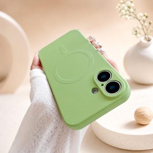 Op de afbeelding: Een lichtgroene smartphonehoes met een cirkelvormige magnetische oplaadring aan de achterkant en een verhoogde cameramodule met twee ronde lensuitsparingen. De hoes heeft een gladde, matte afwerking en een nauwkeurige uitsparing voor de flitser.