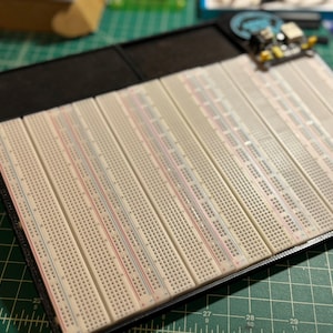 May include: A collection of white breadboards with red, blue, and black lines, arranged in a black frame. A small circuit board with components is placed on top. The breadboards are on a green surface.