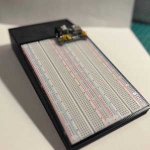 May include: A white breadboard with red and blue lines, set in a black frame. A small circuit board with various components is placed on top. This item is used for electronics projects.