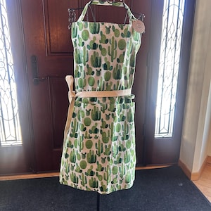 Puede incluir: Delantal con estampado de cactus verde y blanco, exhibido en un soporte de metal negro. El delantal presenta un patrón repetido de cactus verdes sobre un fondo blanco. Tiene una cinta en la cintura y una correa para el cuello de color beige.