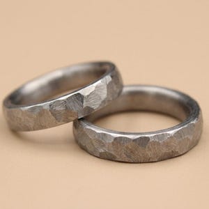 May include: Two silver rings with a hammered texture are shown. The rings are stacked, with one partially covering the other. The background is a plain, light tan colour.