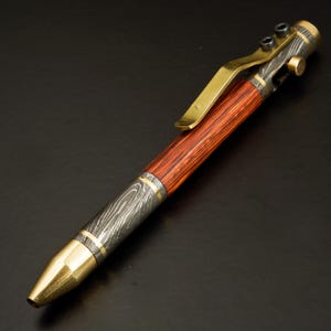 May include: A handcrafted pen with a wooden barrel and metal accents. The pen features a reddish-brown wood grain barrel, a brass clip, and a metal tip. The metal components have a silver and gold finish. The pen is designed for writing.