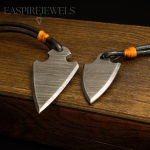 May include: Two matching arrowhead pendants with a textured, metallic finish. Each pendant is suspended from a black leather cord, accented with orange thread. The pendants rest on a dark wooden surface, showcasing the jewelry's craftsmanship.