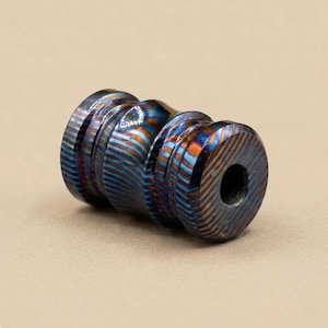 May include: A cylindrical metal bead with a blue and bronze striped pattern. The bead has a central indentation and a hole through one end. The surface has a textured appearance.