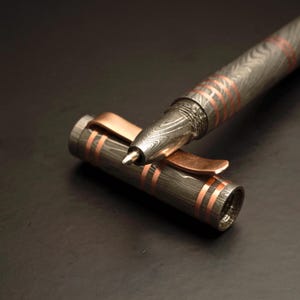 May include: A detailed close-up of a rollerball pen with a Damascus steel barrel and copper accents. The pen's cap is open, revealing the writing tip. The pen has a textured, patterned design with copper-colored bands.