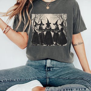 Peut inclure: Un t-shirt gris anthracite à manches courtes avec une impression graphique représentant cinq silhouettes en robes noires et chapeaux pointus debout en cercle dans une forêt. Le texte "Girls Will Be Girls." est imprimé en blanc au centre.