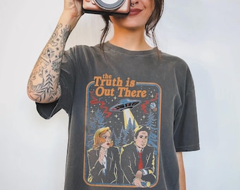 May include: Dark grey t-shirt with the text "the Truth is Out There" and a graphic of a UFO and two people in a forest. The shirt is worn by a person holding a rose gold instant camera.