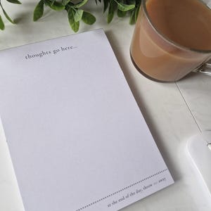 Peut inclure: Un bloc-notes blanc avec le texte "thoughts go here..." et "at the end of the day, throw me away" imprimé en gris. Une tasse en verre transparent remplie d'une boisson brun clair et une souris d'ordinateur blanche sont également visibles.