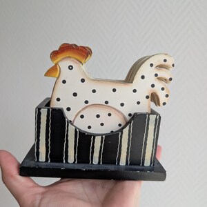 May include: A wooden chicken-shaped coaster set. The chicken is white with black polka dots and a red and yellow comb. The holder is black with white vertical stripes. The set is held in a hand.