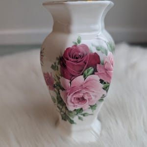 May include: White ceramic vase featuring a floral rose design in pink and burgundy hues. The vase has a flared top with gold trim and a hexagonal base. A decorative item for home interiors.