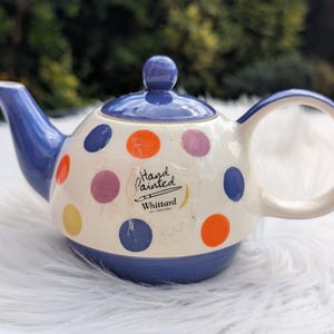 Può includere: Una teiera in ceramica dipinta a mano con base bianca, decorata con pois blu, arancioni, viola e gialli. La teiera ha coperchio, beccuccio e manico blu. Il testo "Hand Painted" e "Whittard of Chelsea" è visibile.
