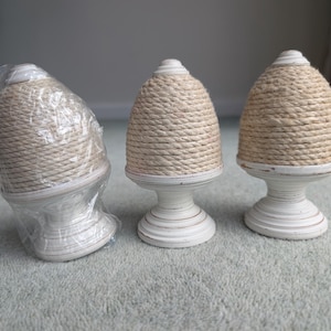 Puede incluir: Tres remates decorativos. Tienen una parte superior beige envuelta en cuerda y una base blanca con forma de pedestal. Uno está envuelto en plástico. Están sobre una superficie gris claro.