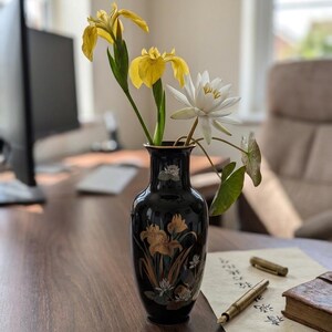 Può includere: Un vaso nero con disegni floreali dorati contiene iris gialli e una ninfea bianca. Il vaso è appoggiato su una scrivania di legno con una penna, carta e un piccolo libro. Lo sfondo include un monitor di computer e una sedia.