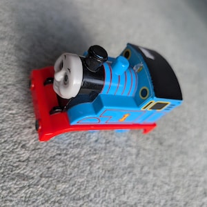 May include: A blue toy train with red accents and the number 1 on the side. The train has a smiling face and a black smokestack. The toy train is a classic children's toy.