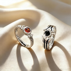May include: Two silver rings with gemstone accents. One ring features a red gemstone, while the other has a black gemstone. The rings are set against a soft, cream-colored background, highlighting their intricate designs and details.
