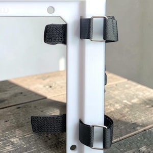 May include: A white rectangular panel with two black fabric straps secured by metal buckles. The straps are attached to the panel with hook and loop fasteners. The panel is on a wooden surface.