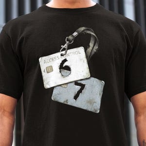 May include: Black t-shirt featuring a graphic of two access control cards with the numbers 6 and 7 cut out. The top card reads "ACCESS CONTROL". The cards are attached to a lanyard with a metal clasp.