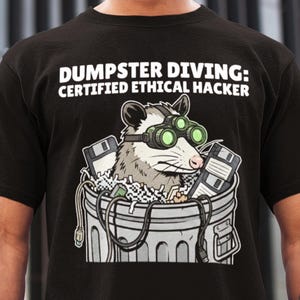 Op de afbeelding: Zwart t-shirt met de tekst "DUMPSTER DIVING: CERTIFIED ETHICAL HACKER". De afbeelding toont een wasbeer met een nachtkijker in een vuilnisbak vol diskettes en computeronderdelen.