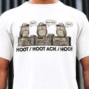 May include: White t-shirt with a cartoon illustration of three owls wearing caps and perched on electronic devices. Speech bubbles above the owls read "Hoot" and "Hoot-ACK." The text "HOOT / HOOT ACK / HOOT" is printed below the owls.