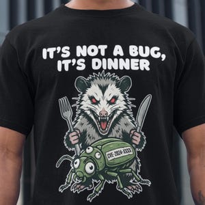 Peut inclure: T-shirt noir avec un dessin d'opossum tenant une fourchette et un couteau, avec un insecte vert en dessous. Le texte au-dessus dit "IT'S NOT A BUG, IT'S DINNER". L'insecte porte le texte "CVE-2024-XXXX".