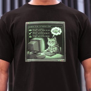 Op de afbeelding: Zwart t-shirt met een afbeelding van een kat met een bril en een hoodie, die op een computer werkt. Het scherm toont code en een denkbeeldige wolk met een vis. Het ontwerp heeft een retro, pixelachtige uitstraling.