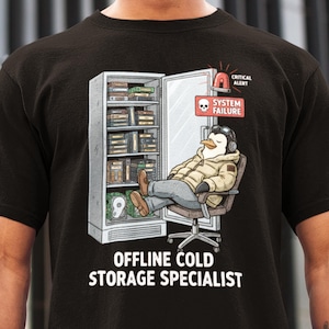 Op de afbeelding: Zwart t-shirt met een cartoonpinguïn met een headset en jas, zittend in een bureaustoel voor een koelopslag. De tekst "OFFLINE COLD STORAGE SPECIALIST" staat onder de afbeelding, met een "SYSTEM FAILURE"-waarschuwing.