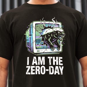 May include: Black t-shirt featuring a graphic of a badger bursting through a retro computer monitor. The monitor is outlined in purple and green, with the text "I AM THE ZERO-DAY" in white below the graphic.