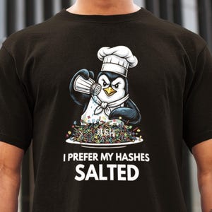 May include: Black t-shirt with a cartoon penguin chef salting a plate of food. The penguin wears a chef's hat and apron. Text on the shirt reads "I PREFER MY HASHES SALTED" in white. The food is colorful.