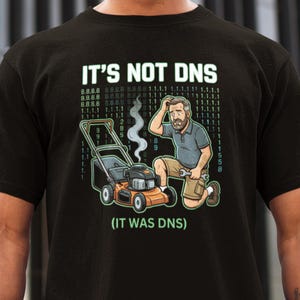 May include: Black t-shirt featuring the text "IT'S NOT DNS" above a cartoon of a lawnmower and a man. The lawnmower is orange and black, and the man is wearing a blue shirt and khaki shorts. The text "(IT WAS DNS)" is below.